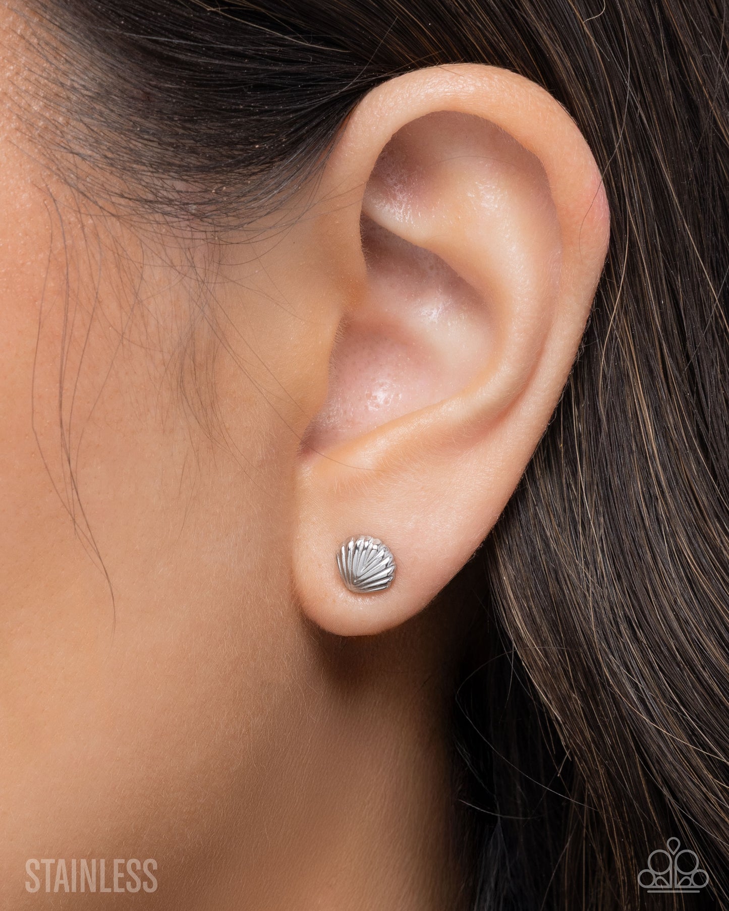 Close-up of an ear wearing a silver shell stud earring with blurred hair and skin in the background.