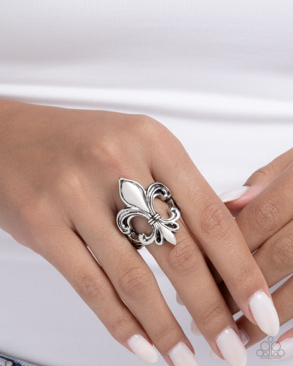 Silver fleur-de-lis ring on a hand with light pink nail polish against a white background