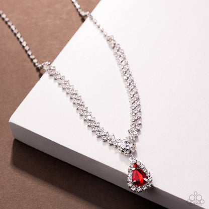 A silver-tone double-strand necklace with clear crystals and a teardrop-shaped red gemstone pendant, displayed on a white rectangular block against a brown background.