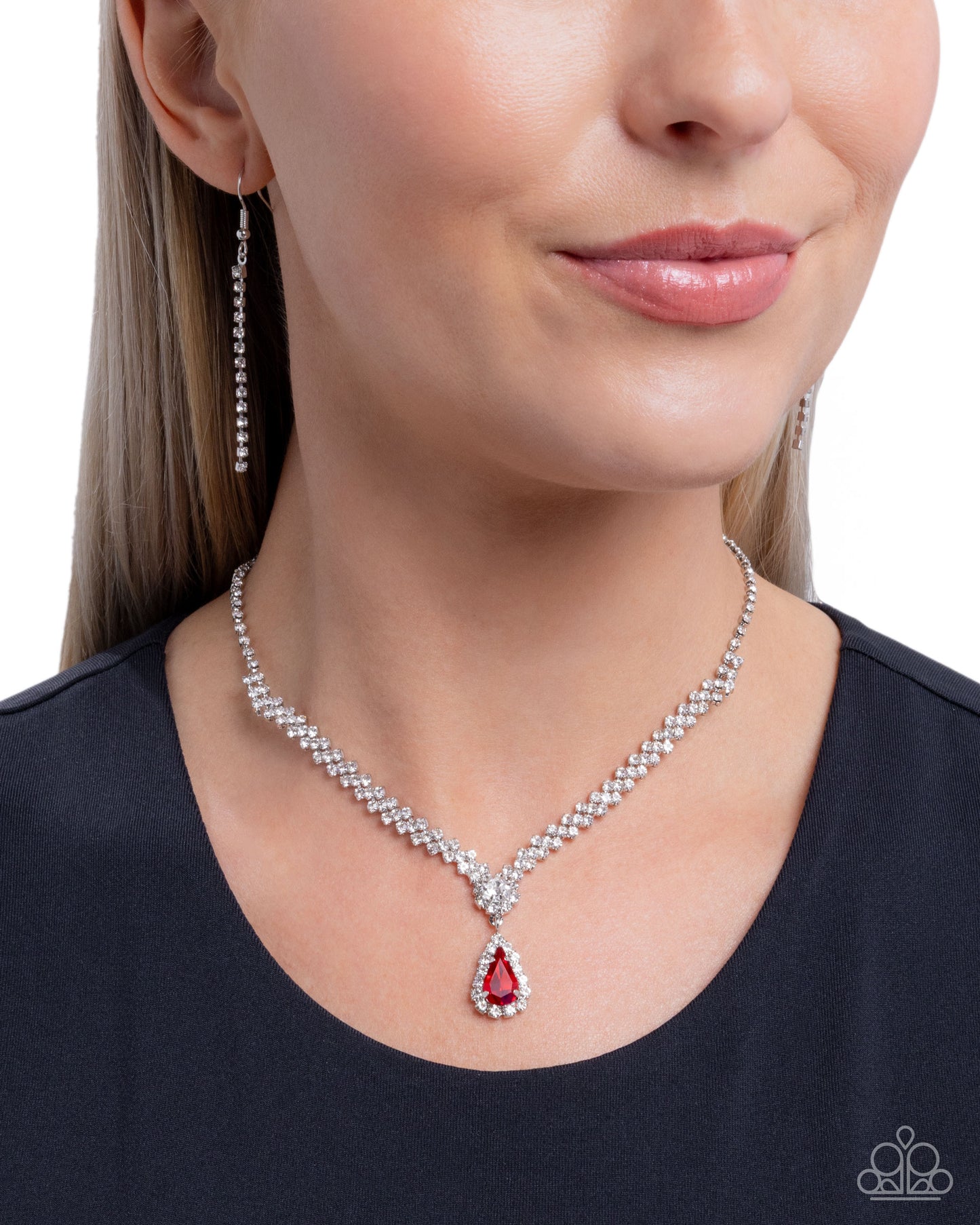 A close-up of a woman wearing a silver-tone necklace with a teardrop-shaped red gemstone pendant, matching drop earrings, and a black top against a white background.