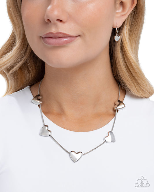 A close-up of a woman wearing a silver necklace with five polished heart charms spaced along a delicate chain, paired with a white top against a clean white background.