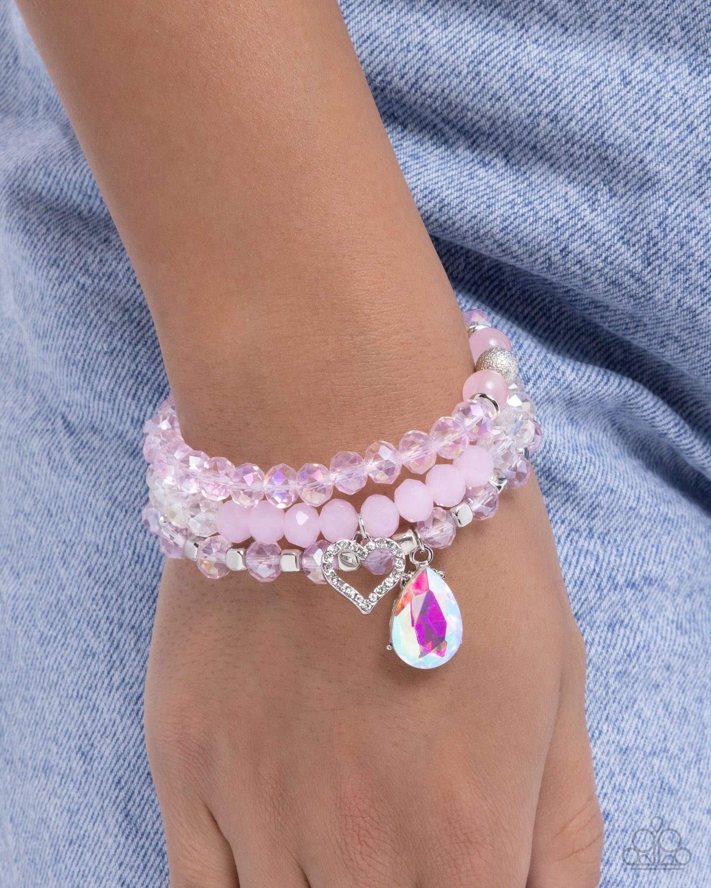 Pink beaded bracelet with a heart and teardrop charm on a wrist against a denim background