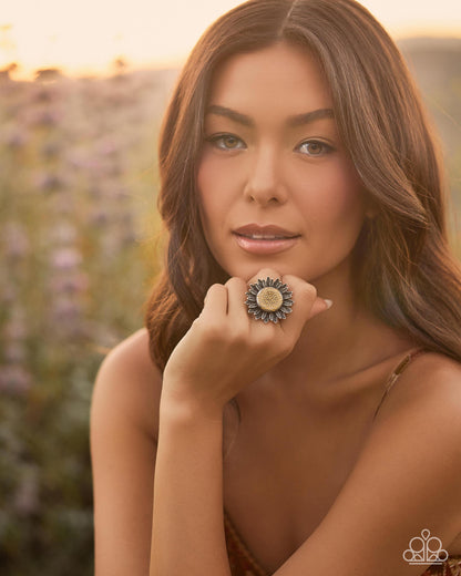 A woman in a warm outdoor setting wearing a large, ornate ring with a sunflower-like design featuring blue, gold, and cream tones.