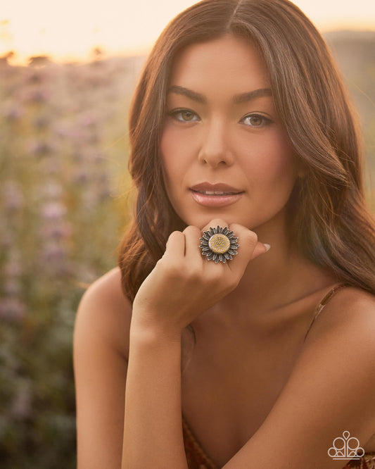 A woman in a warm outdoor setting wearing a large, ornate ring with a sunflower-like design featuring blue, gold, and cream tones.