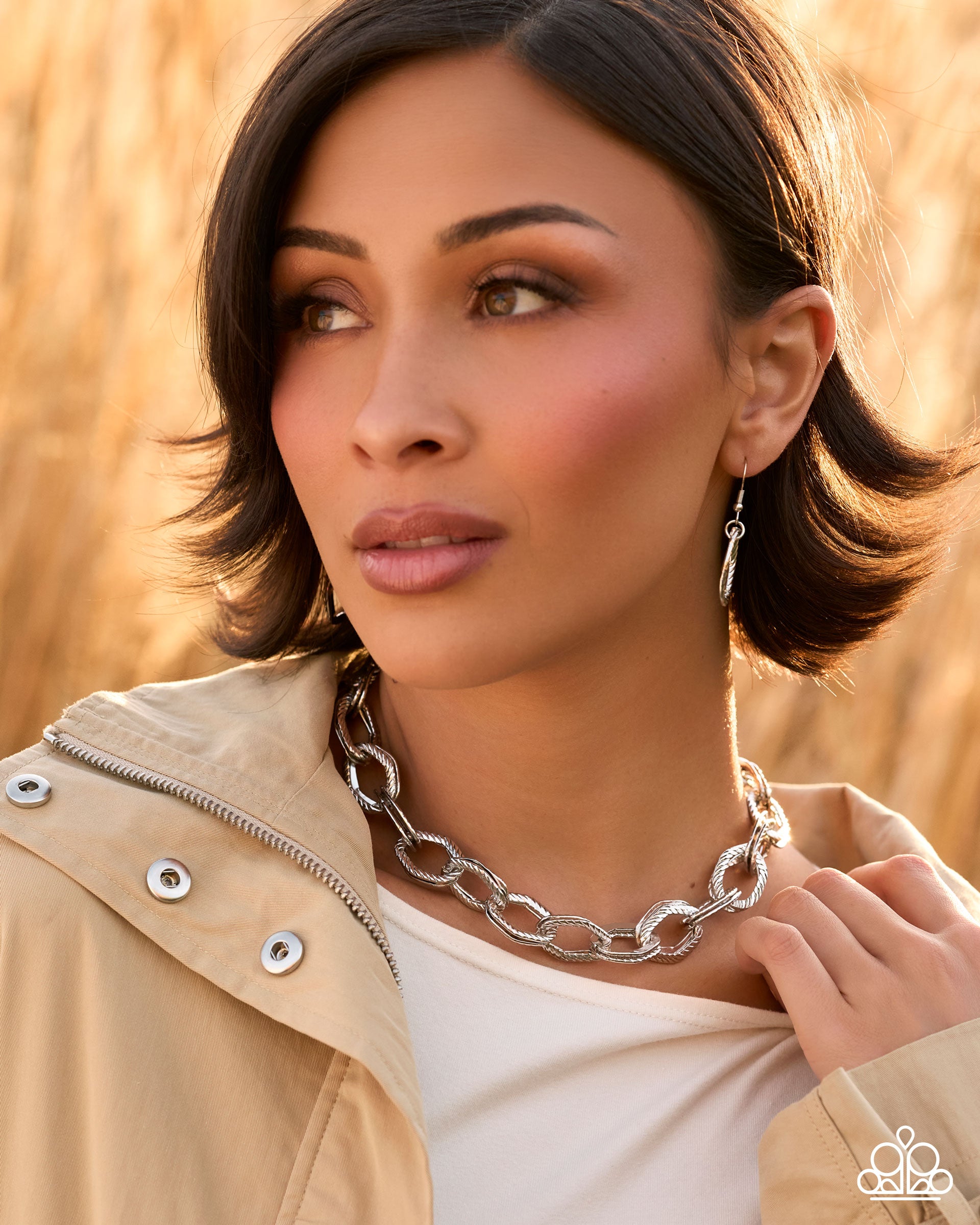 A woman with short dark hair wearing a chunky silver chain necklace and matching drop earrings, photographed outdoors against a warm golden field background.