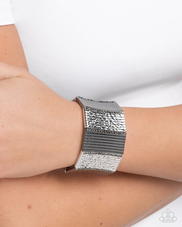 A close-up of a person's wrist wearing a wide silver cuff bracelet with textured and mesh-like bands, against a white top background.