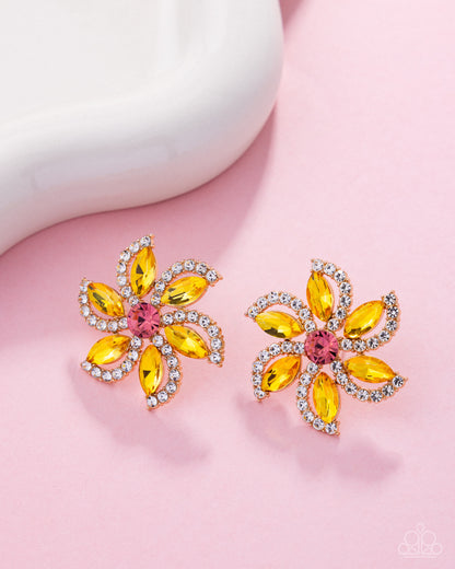 A pair of flower-shaped stud earrings with yellow marquise-cut stones, a pink round center stone, and clear rhinestone borders, displayed on a pink surface with a white curved prop.