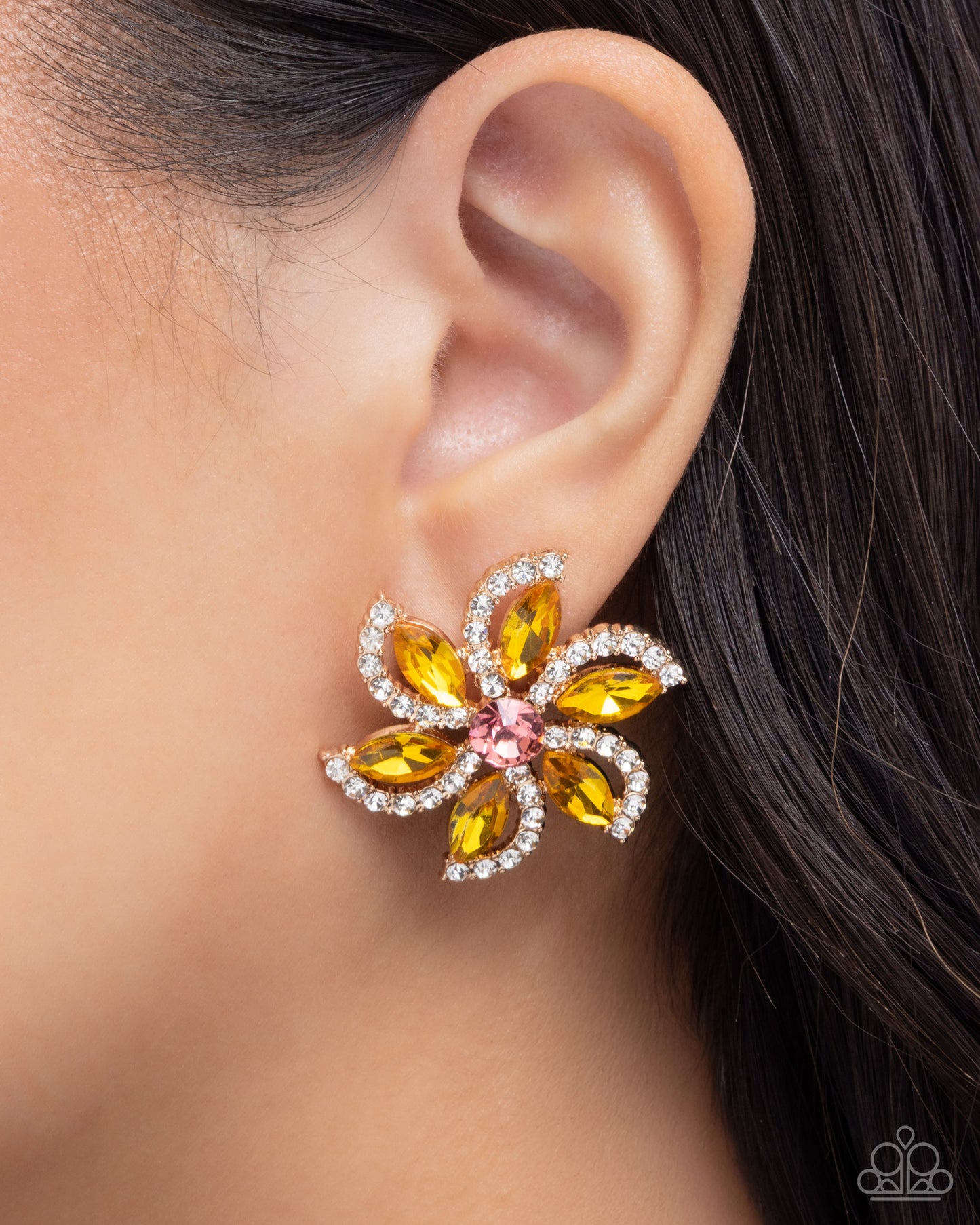A close-up of a model's ear wearing a flower-shaped earring with yellow marquise-cut crystals, a pink center stone, and clear rhinestone borders.