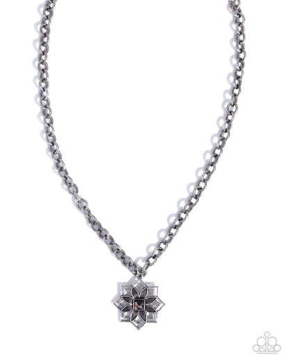 A silver-toned chain necklace with a geometric starburst pendant featuring clear rhinestones and a small dark center, shown on a plain white background. A Paparazzi logo watermark is visible in the bottom right corner.