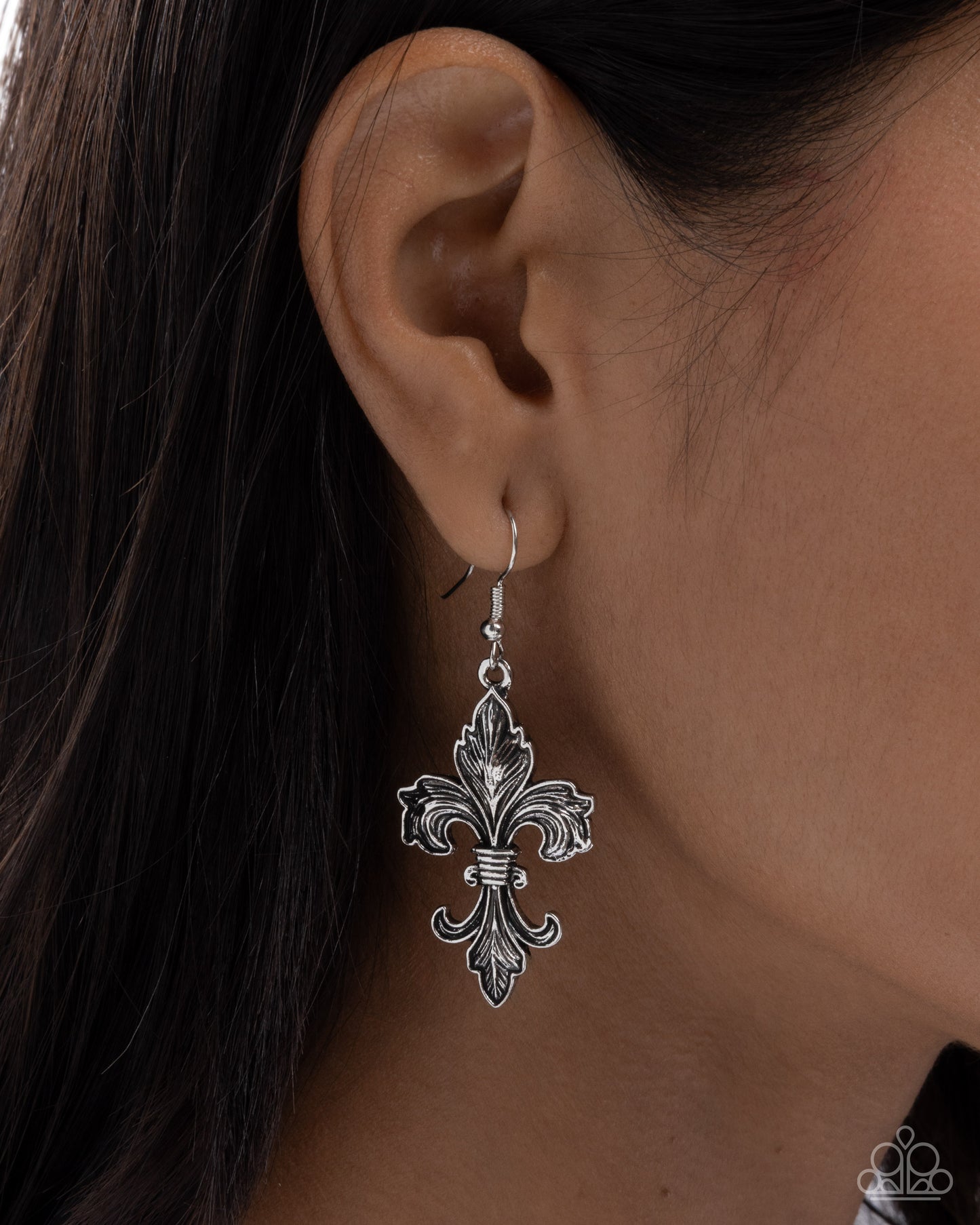 Silver fleur-de-lis earring worn by a person with dark hair.