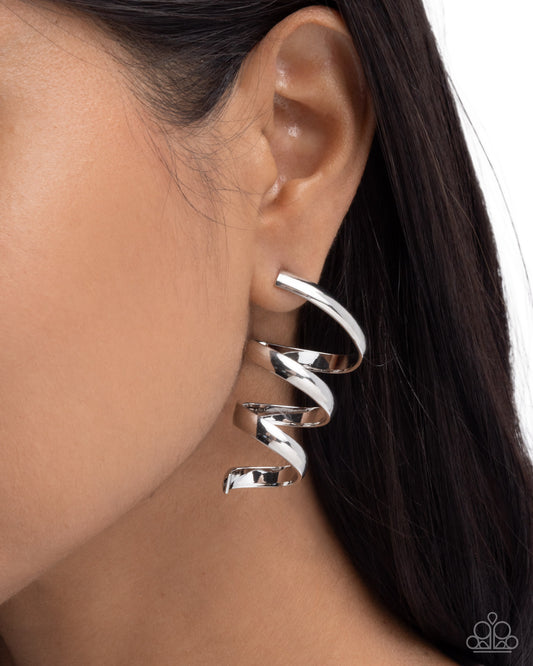 A close-up of a person's ear wearing a shiny silver spiral hoop earring, with dark hair and a white background.