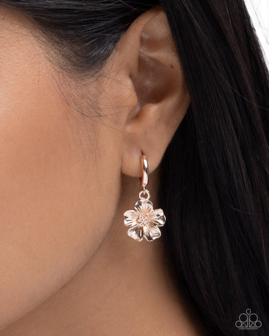 A close-up of a person's ear wearing a rose gold flower-shaped dangle earring with a small clear crystal center.