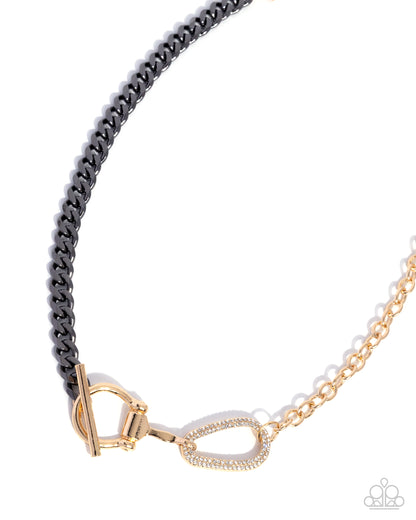 A close-up of a necklace featuring a black curb chain section and a gold chain section, joined by a gold clasp and a small gold heart-shaped charm with a clear stone border, on a white background.