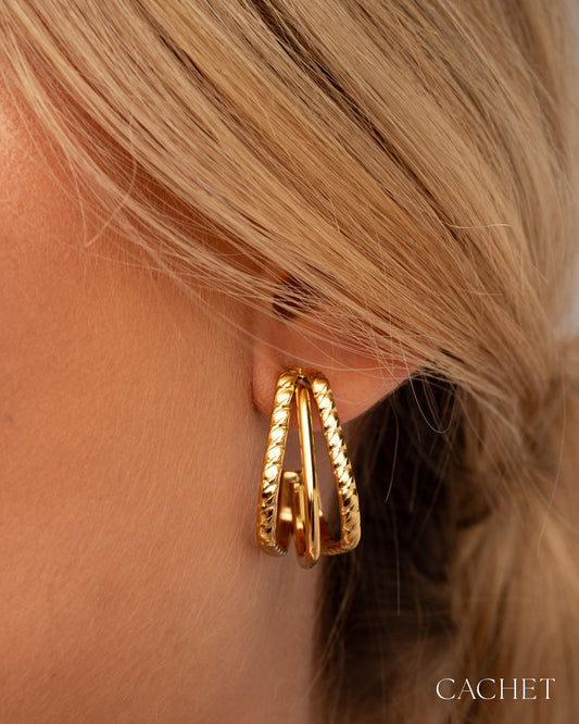 Gold hoop earrings worn by a person with blonde hair, brand 'Cachet' visible.