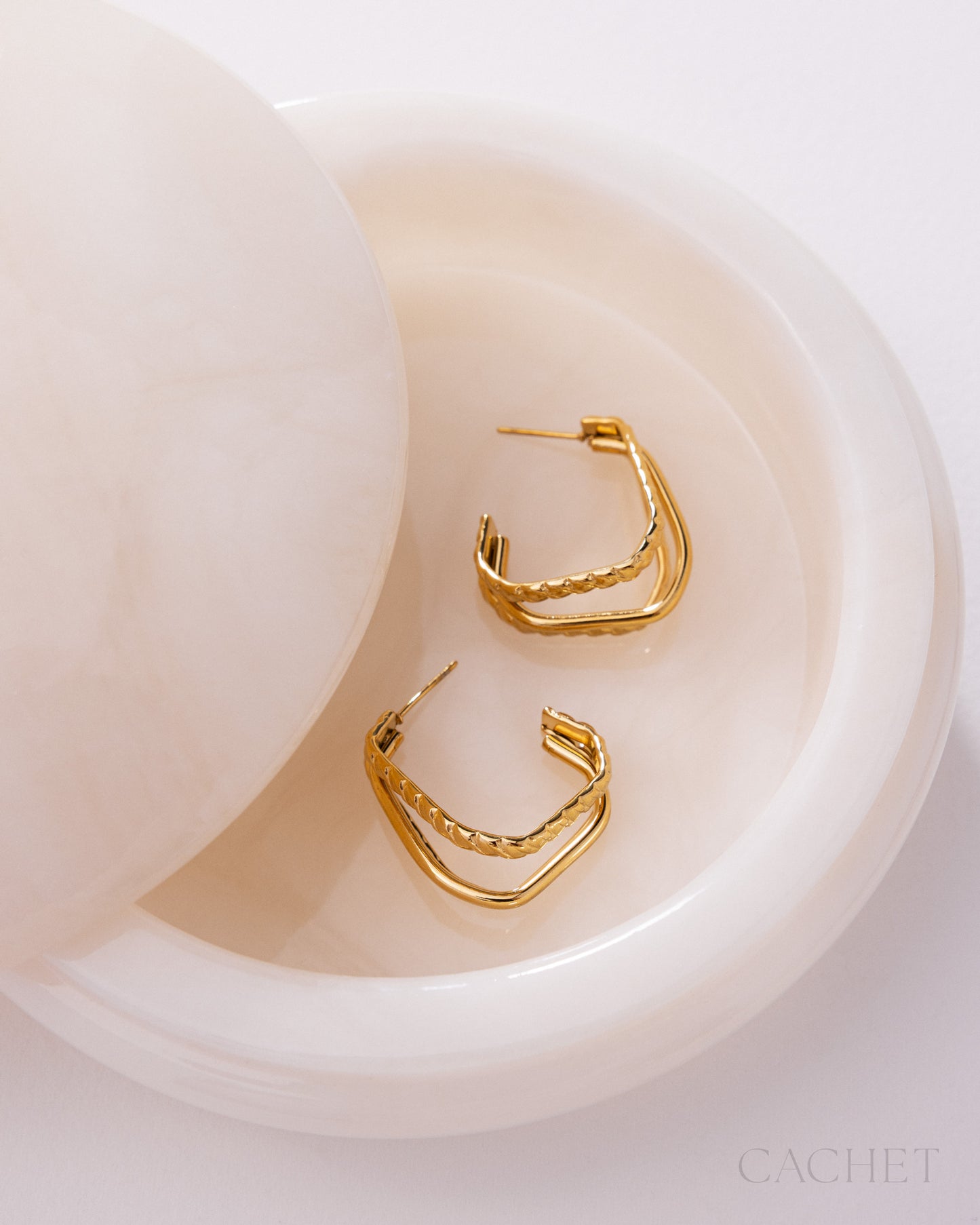 Gold hoop earrings on a white dish with 'Cachet' branding.