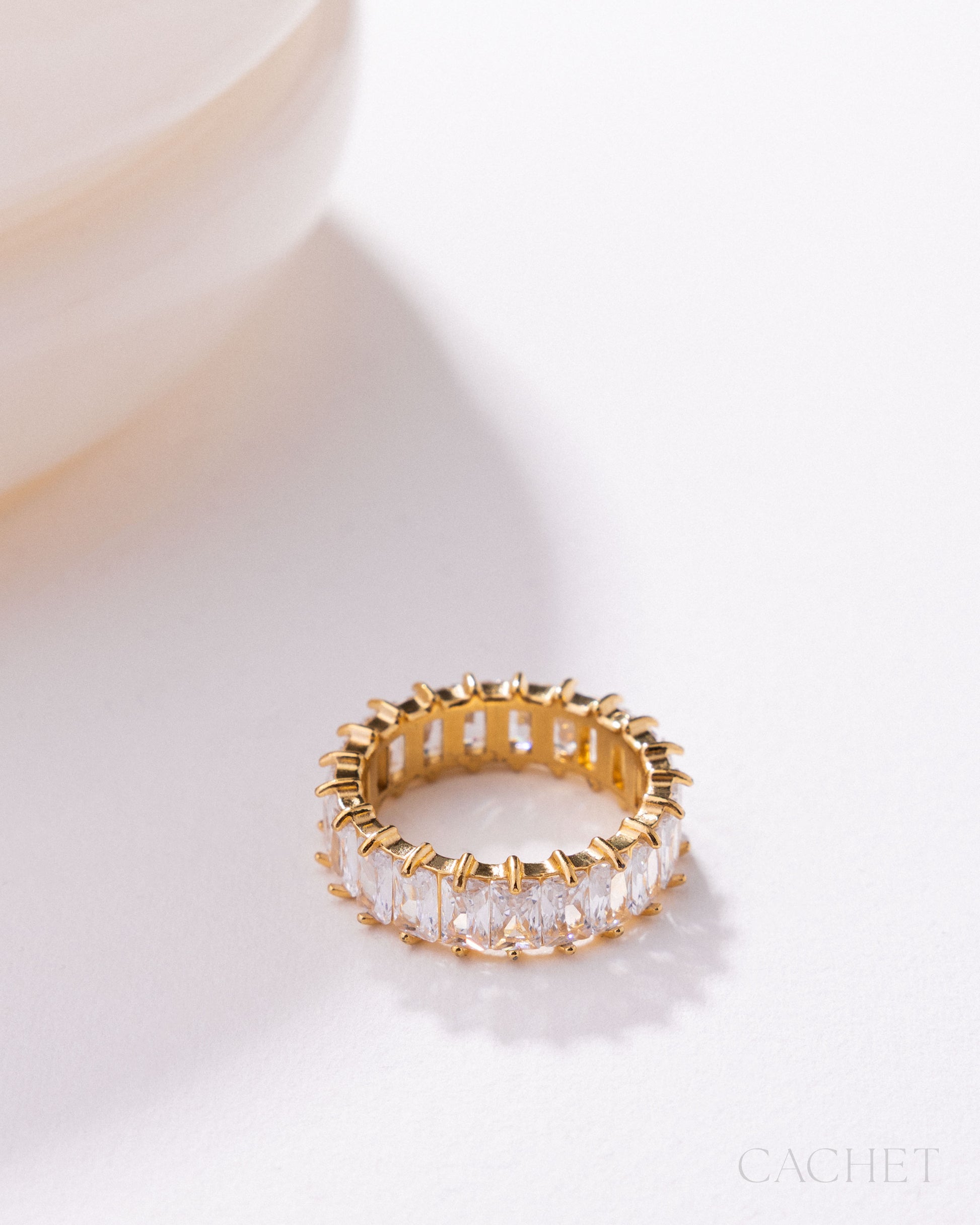 Gold ring with clear gemstones on a light background, featuring the brand 'Cachet'.