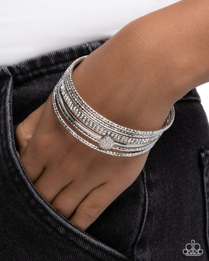 A close-up of a person's hand and wrist wearing a stack of silver-toned bangles, some adorned with clear pavé stones and one with a round pavé centerpiece. The hand is resting in the pocket of dark denim jeans against a clean white background.
