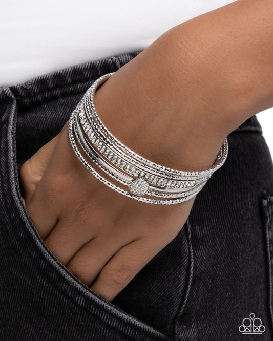 A close-up of a person's hand and wrist wearing a stack of silver-toned bangles, some adorned with clear pavé stones and one with a round pavé centerpiece. The hand is resting in the pocket of dark denim jeans against a clean white background.