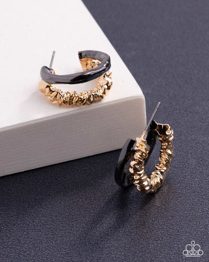 A pair of gold hoop earrings with a textured, ridged outer band and a smooth black inner band, displayed on a white block and a dark gray surface.