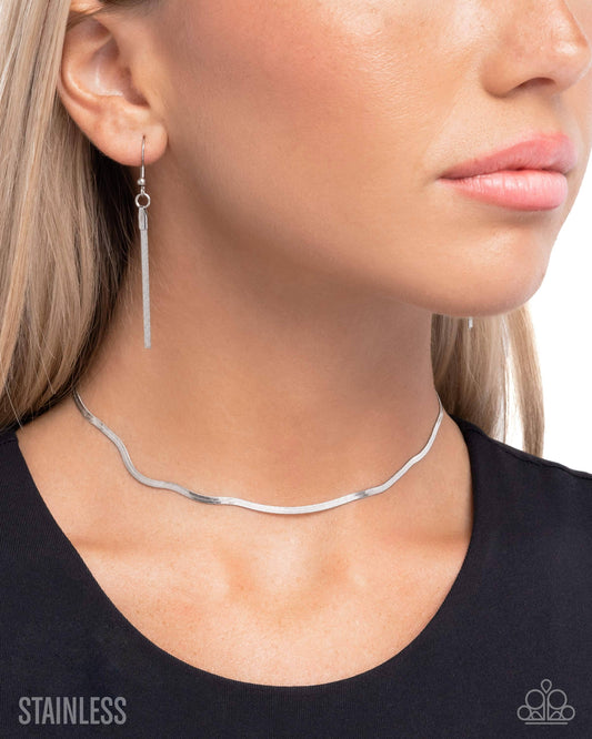 A close-up of a woman wearing a slim stainless steel wavy choker necklace and a matching long drop earring. The image features a 'STAINLESS' text overlay on the bottom left and a small brand logo on the bottom right.