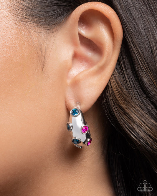 Close-up of an ear wearing a silver hoop earring with colorful gemstones.