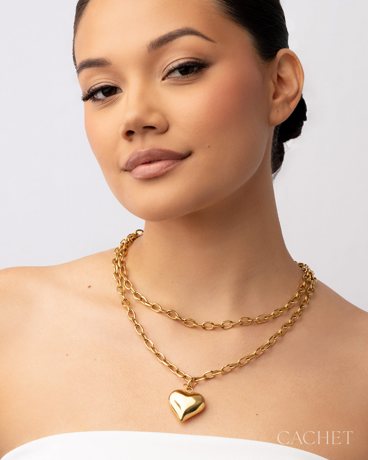 Woman wearing a gold chain necklace with a heart pendant on a plain background