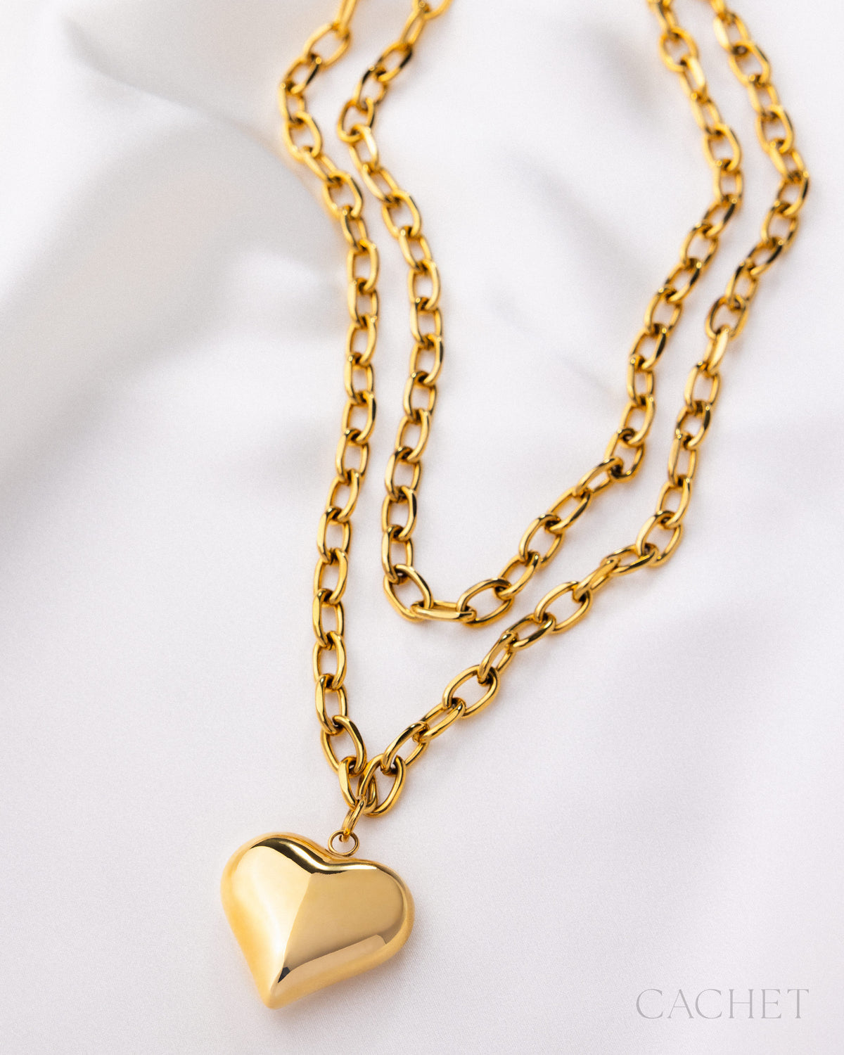 Gold chain necklace with a heart pendant on a white background, featuring the brand 'Cachet'.