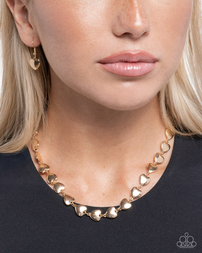 Close-up of a person wearing a gold heart-shaped necklace on a plain background