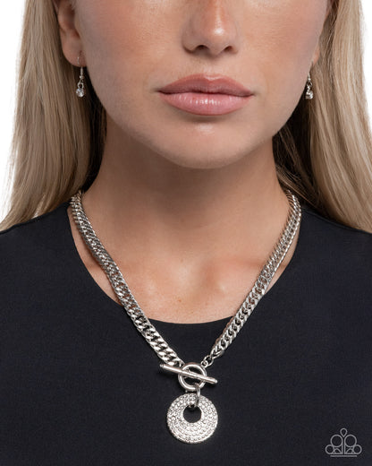 Close-up of a person wearing a silver chain necklace with a circular pendant on a plain background