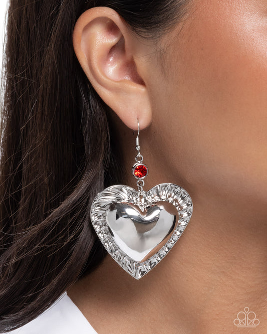 Silver heart-shaped earring with a red gemstone worn by a person.