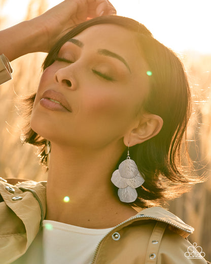 A woman outdoors in warm golden-hour light wearing a pair of large, layered white flower-shaped dangle earrings. She has her eyes closed and one hand near her hair, wearing a beige jacket over a white top.