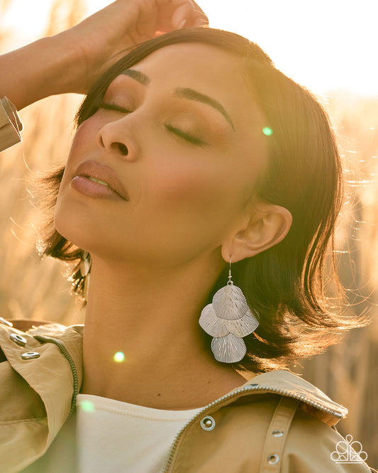 A woman outdoors in warm golden-hour light wearing a pair of large, layered white flower-shaped dangle earrings. She has her eyes closed and one hand near her hair, wearing a beige jacket over a white top.