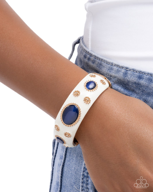 White leather bracelet with blue stones and gold accents on a wrist, against a white background.
