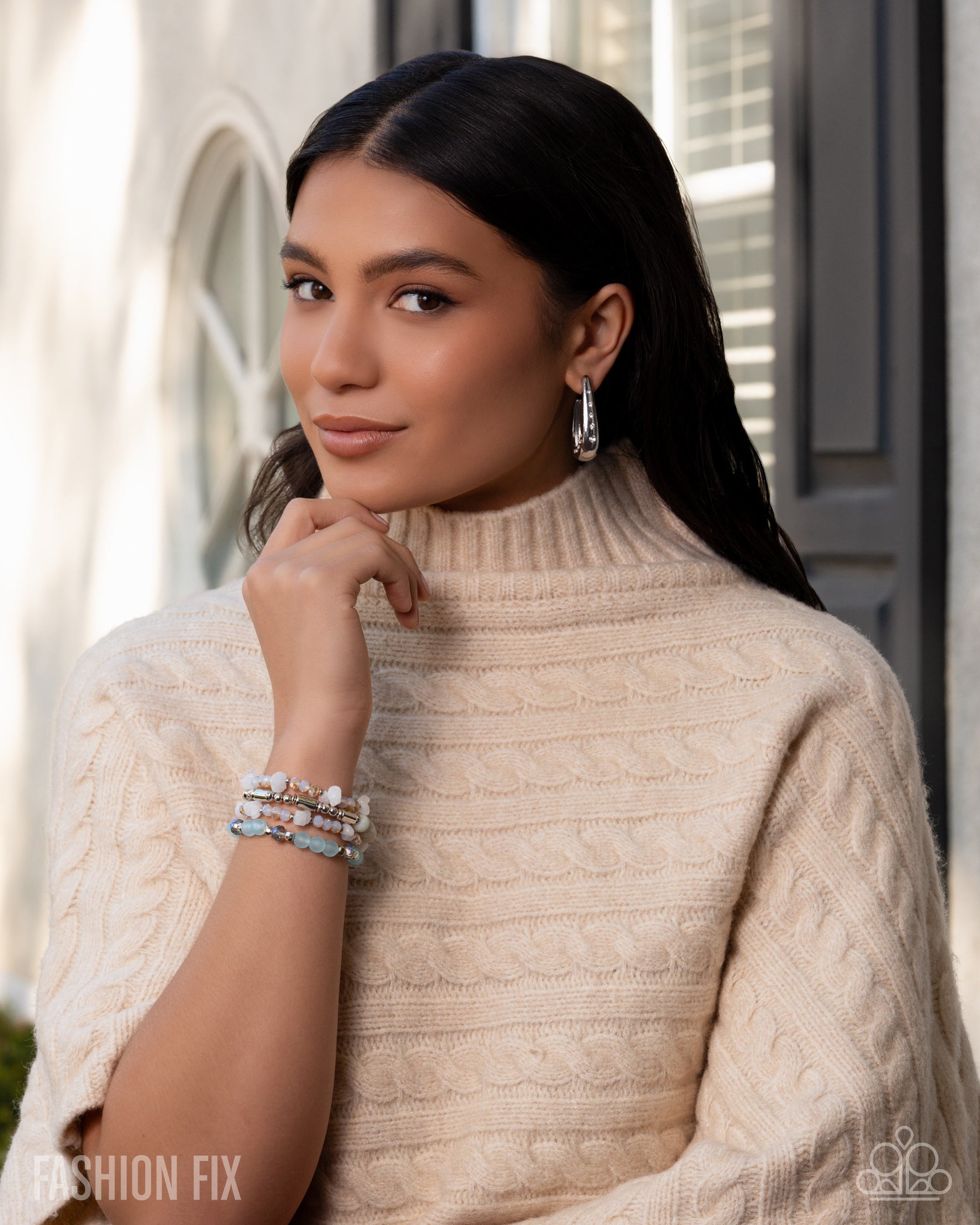 Woman wearing a beige cable knit sweater with jewelry, standing outdoors.