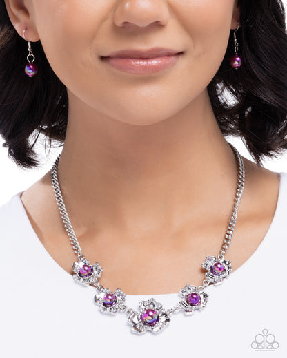 A close-up of a woman wearing a silver-toned statement necklace with five flower-shaped clusters, each set with a purple gemstone and surrounded by clear crystals. Matching purple gemstone drop earrings are also visible.