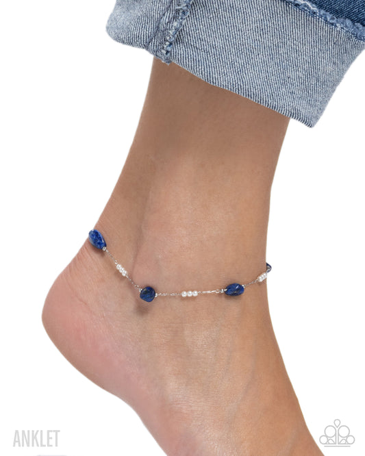 Anklet with blue beads on a foot wearing denim jeans against a white background