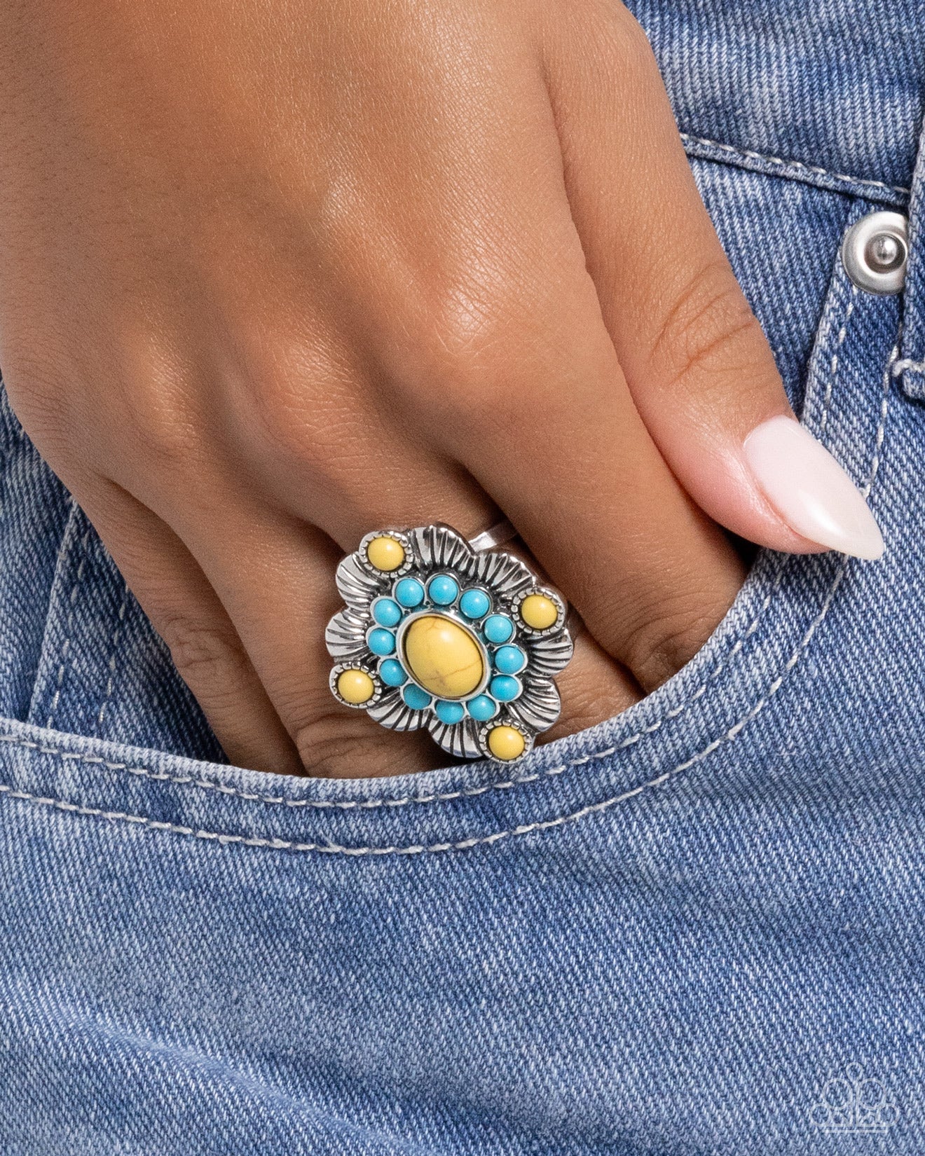Close-up of a hand wearing a decorative ring with turquoise and yellow stones on denim fabric.