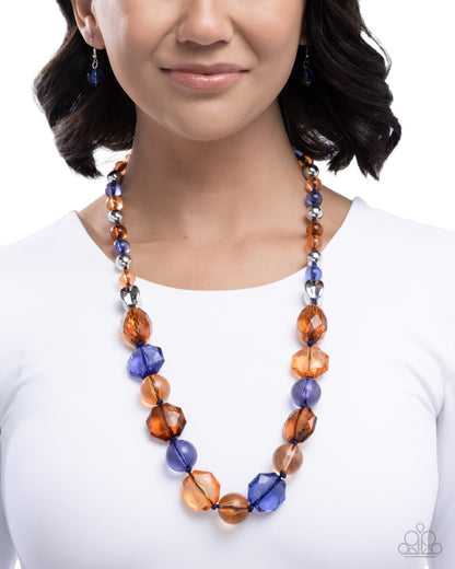 A woman wearing a beaded statement necklace with faceted amber-orange and blue beads, paired with matching blue drop earrings, shown against a white background.