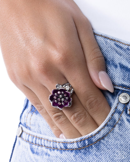 A silver-toned flower-shaped ring with deep purple gemstones and small clear crystals, worn on a finger resting in a pocket of light blue denim jeans.