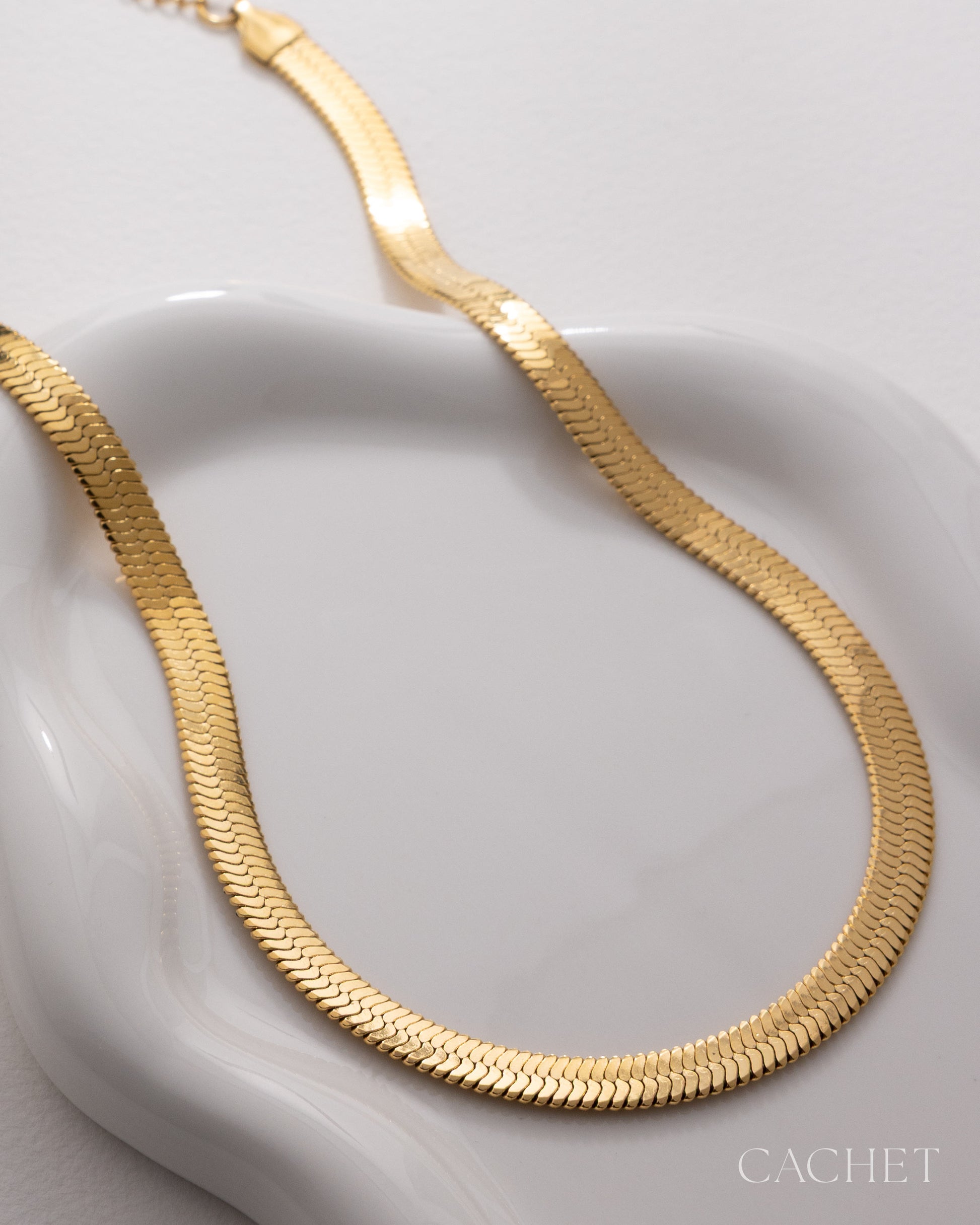 Gold necklace on a white dish with 'Cachet' branding.