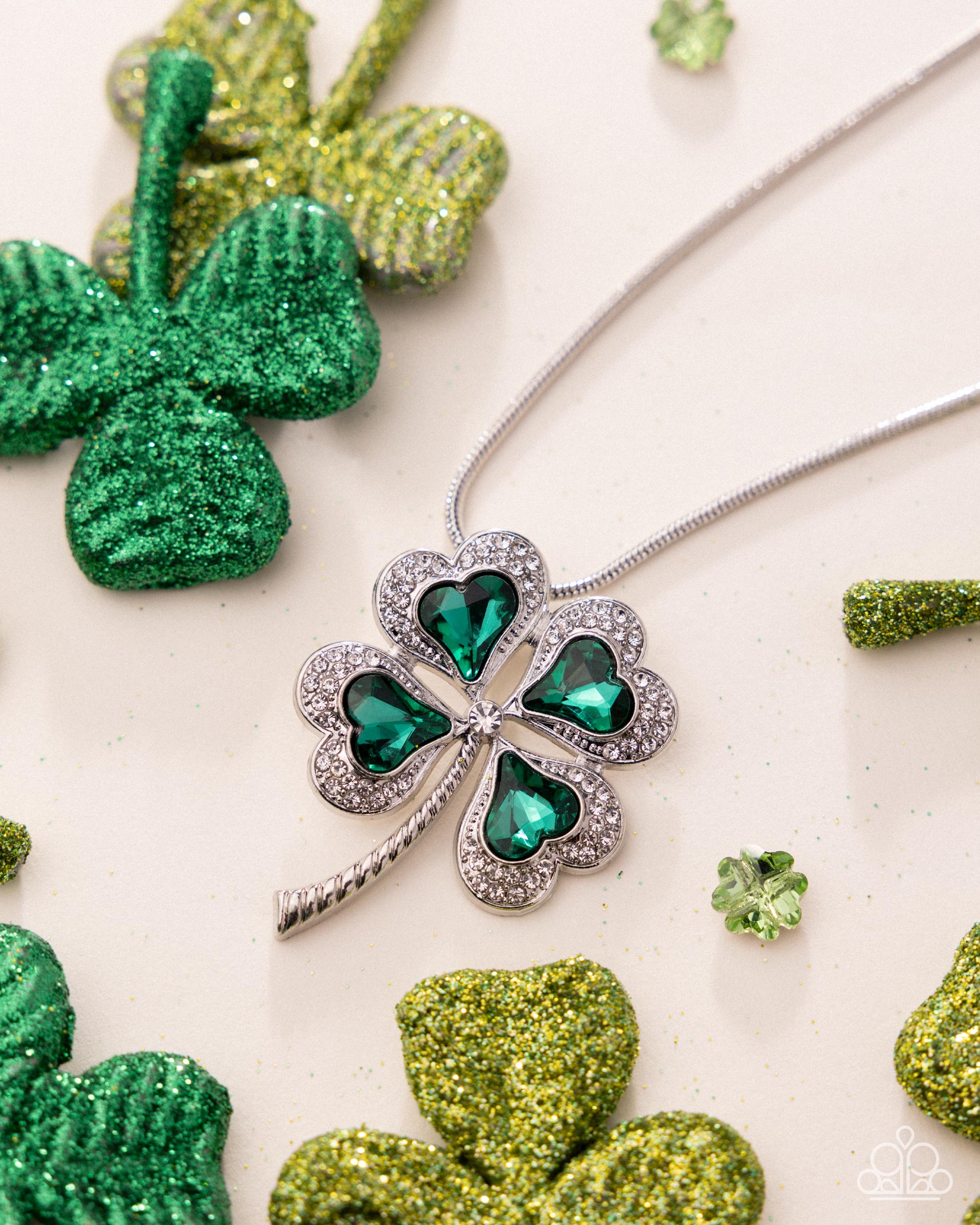 Four-leaf clover necklace with green gemstones on a white background with decorative shamrocks.