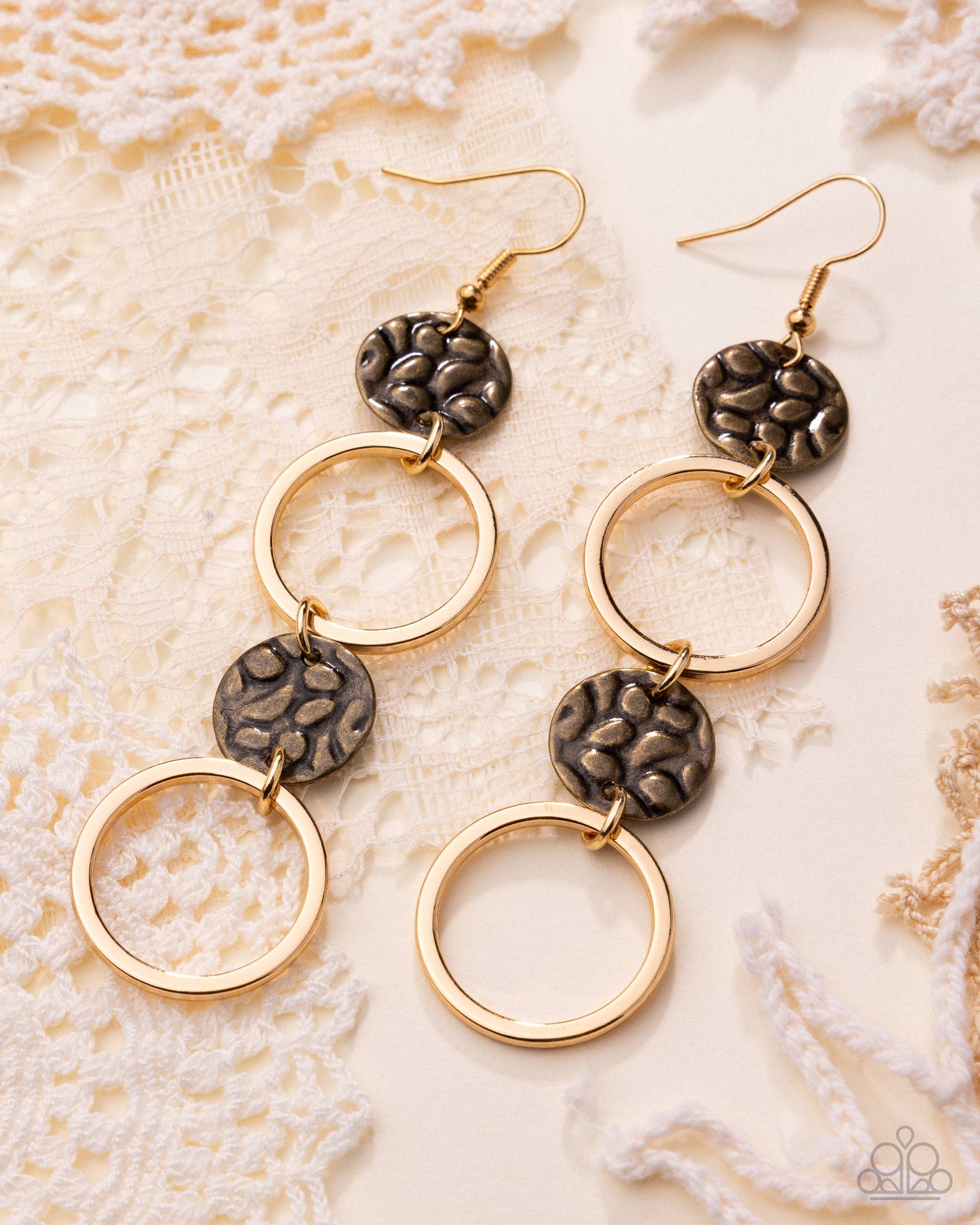 A pair of gold-tone drop earrings with textured dark bronze circular charms and open circular rings, displayed on a cream lace background.