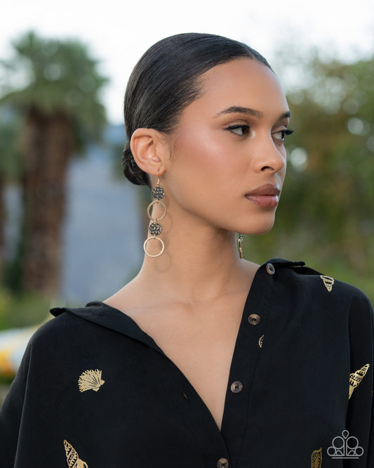 A woman in profile wearing long, dangling gold-toned earrings with circular and filigree details, paired with a black embroidered top, set against a softly blurred outdoor background.