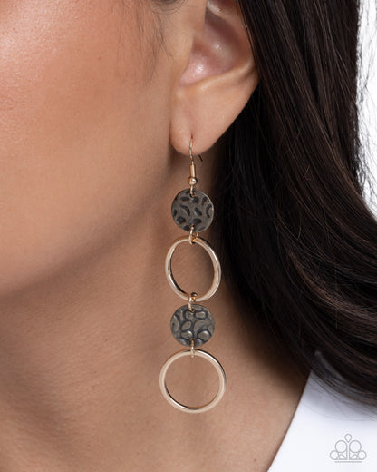 A close-up of a model's ear wearing a three-tier dangle earring. The earring features textured dark gray circular discs and polished gold-tone metal rings.