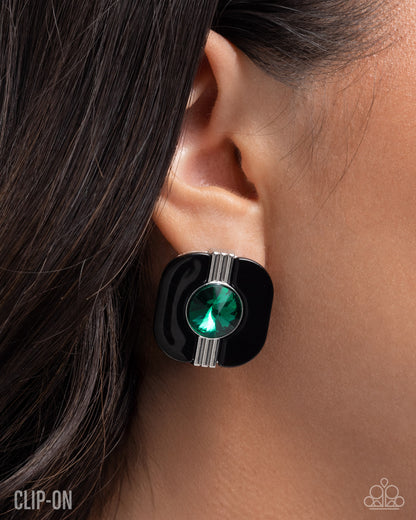 A close-up of a person's ear wearing a black square stud earring with a central green faceted gemstone and silver-toned accent lines.