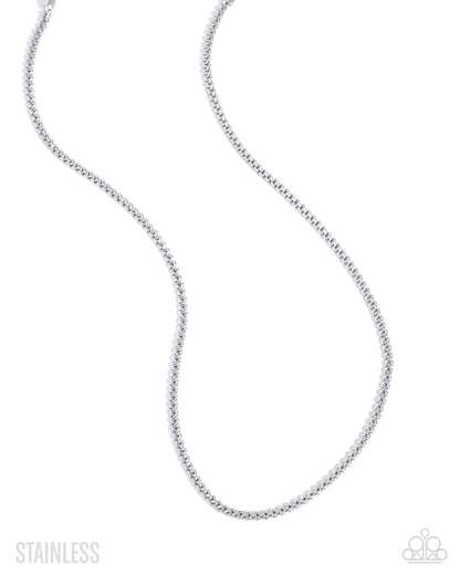 A silver stainless steel box chain necklace, shown in a curved loop shape against a plain white background, with 'STAINLESS' text in the bottom-left corner and a small logo in the bottom-right.