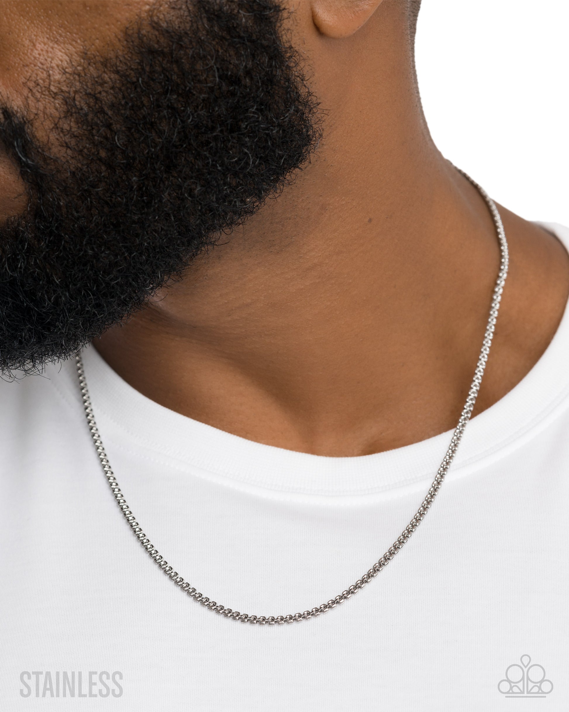 A close-up of a man's neck and beard wearing a silver stainless steel chain necklace, paired with a white t-shirt. The image features a 'STAINLESS' text overlay on the bottom left and a brand logo watermark on the bottom right.