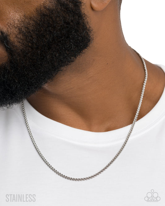 A close-up of a man's neck and beard wearing a silver stainless steel chain necklace, paired with a white t-shirt. The image features a 'STAINLESS' text overlay on the bottom left and a brand logo watermark on the bottom right.
