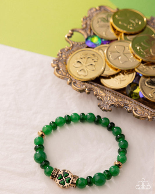 Green beaded bracelet with clover charm on a white surface with gold coins in the background
