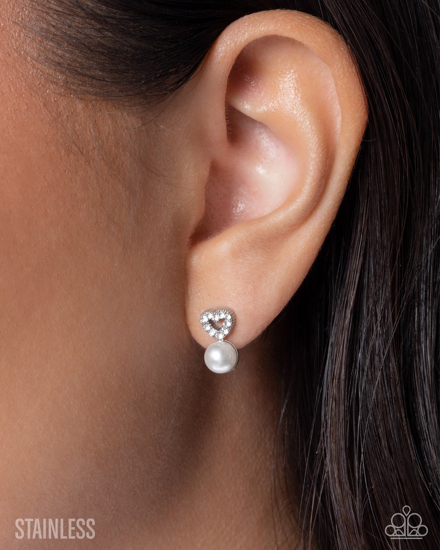 A close-up of a person's ear wearing a small silver stud earring with a heart-shaped clear crystal top and a dangling white pearl. The word 'STAINLESS' appears in the lower left corner.